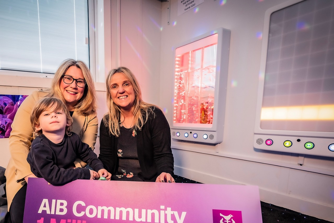 Two adults and a child in a sensory room with banner saying 2024 AIB Community 1 Million Fund Charities!