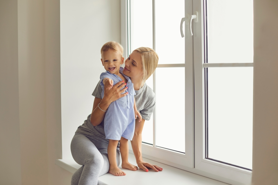 Woman holding her toddler near by a window in their home.