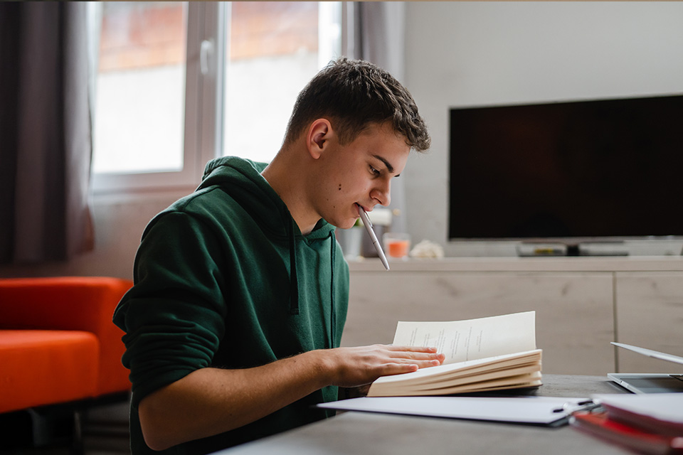 A boy in a green hoodie sitting at his desk reading a book